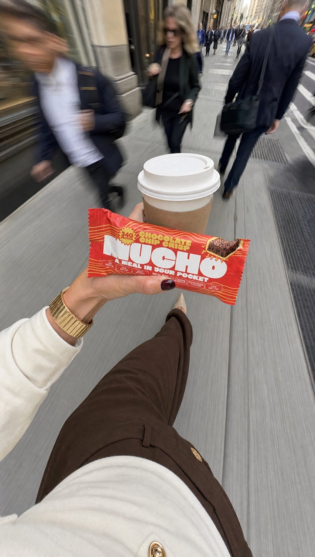 First-person view walking down a busy city street: a hand holding a Chocolate Chip Crisp Mucho bar and a takeaway coffee, commuters blurred past on either side.