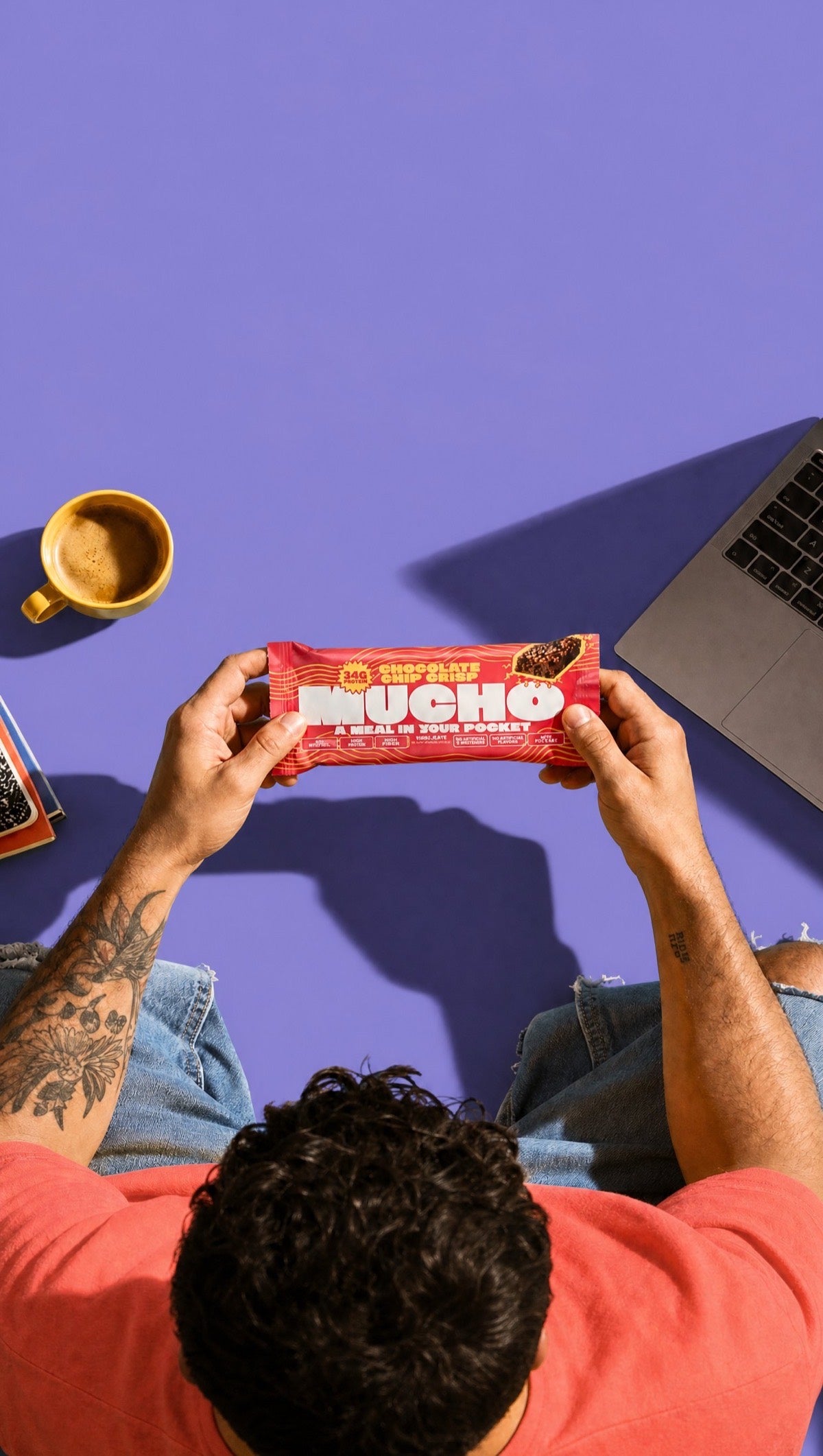 Overhead shot of a man holding a Mucho Chocolate Chip Crisp bar in both hands against a periwinkle background with coffee and laptop